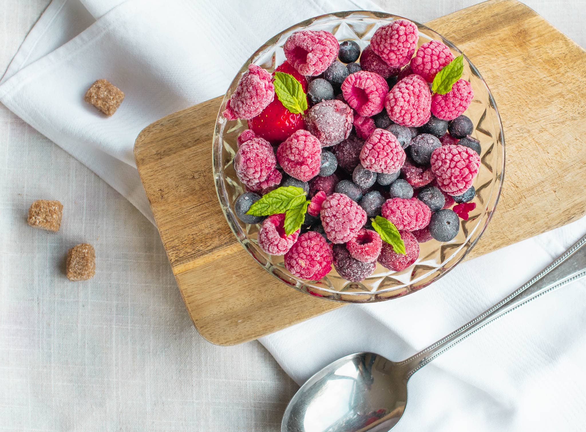 Glass bowl of frozen berries on wooden board with metal spoon and sugar cubes