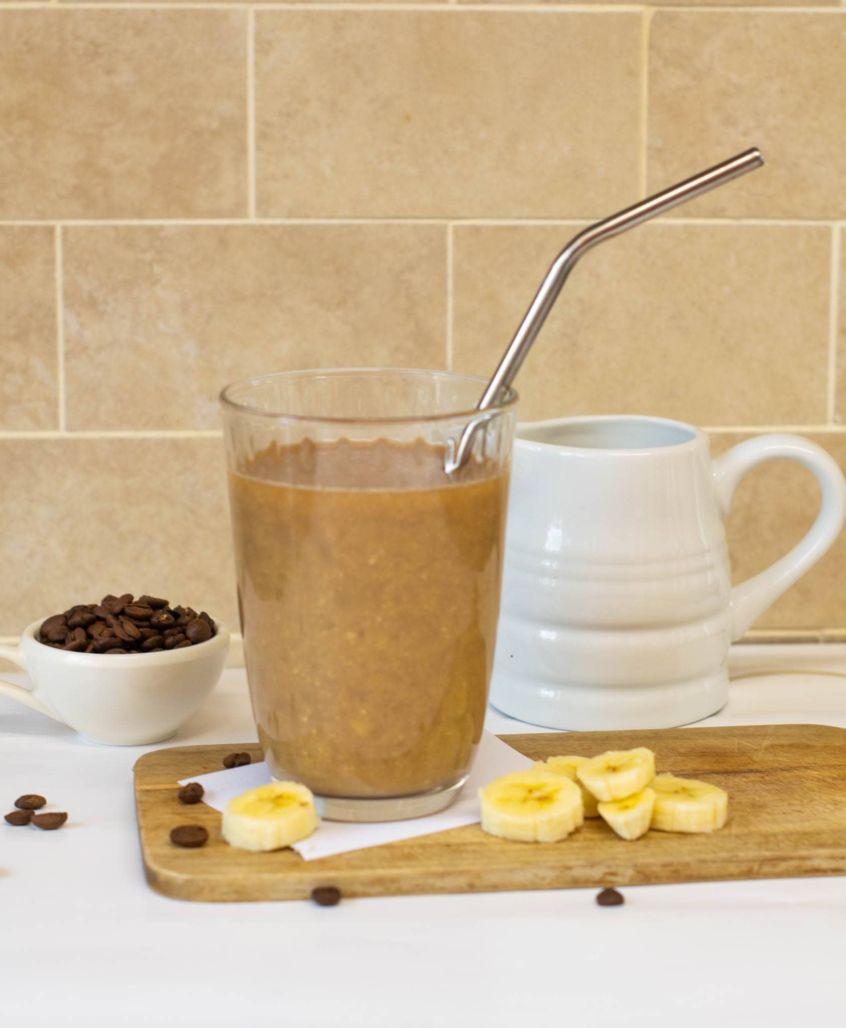 Coffee banana smoothie with metal straw sitting on chopping board with milk jug, coffee beans and banana slices