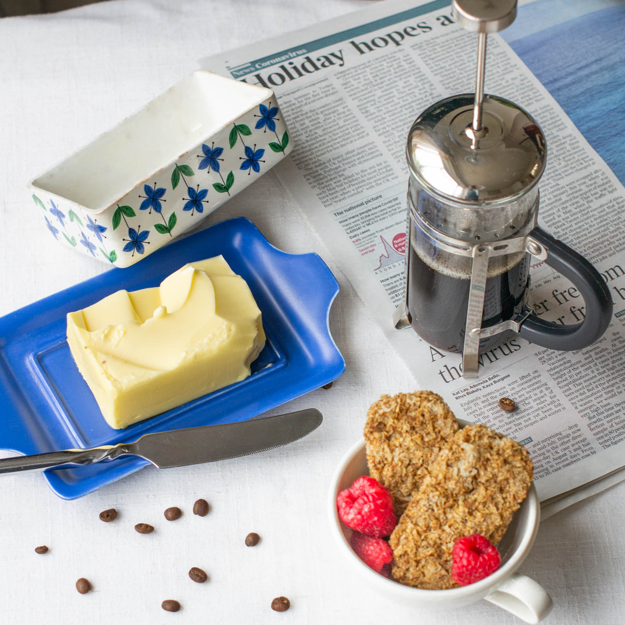 Mini cafetiere sitting on open copy of The Times beside blue butter dish and bowl of Weetabix with raspberries