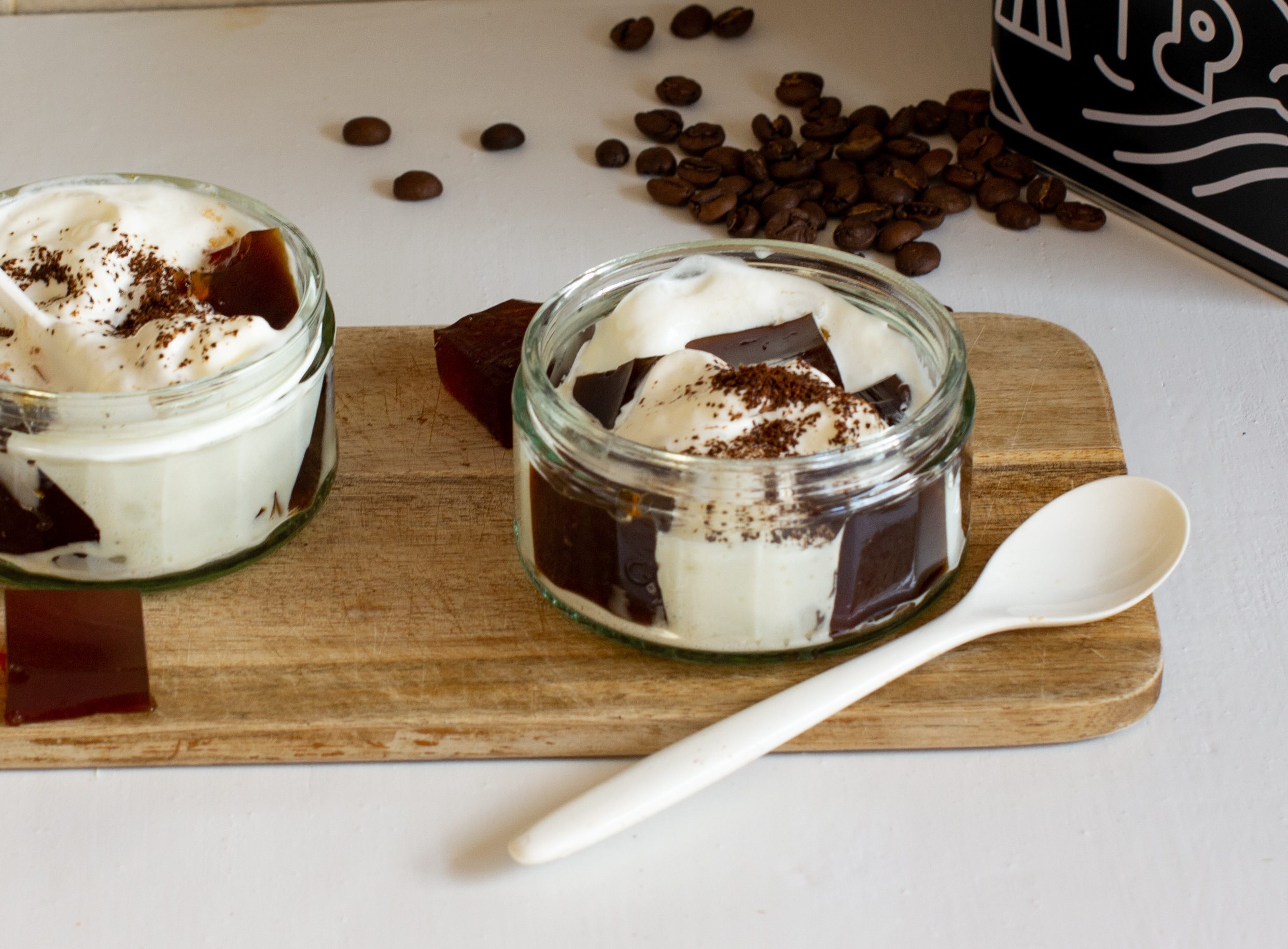 Two glass ramekins of coffee jelly and cream on a chopping board with plastic spoon 
