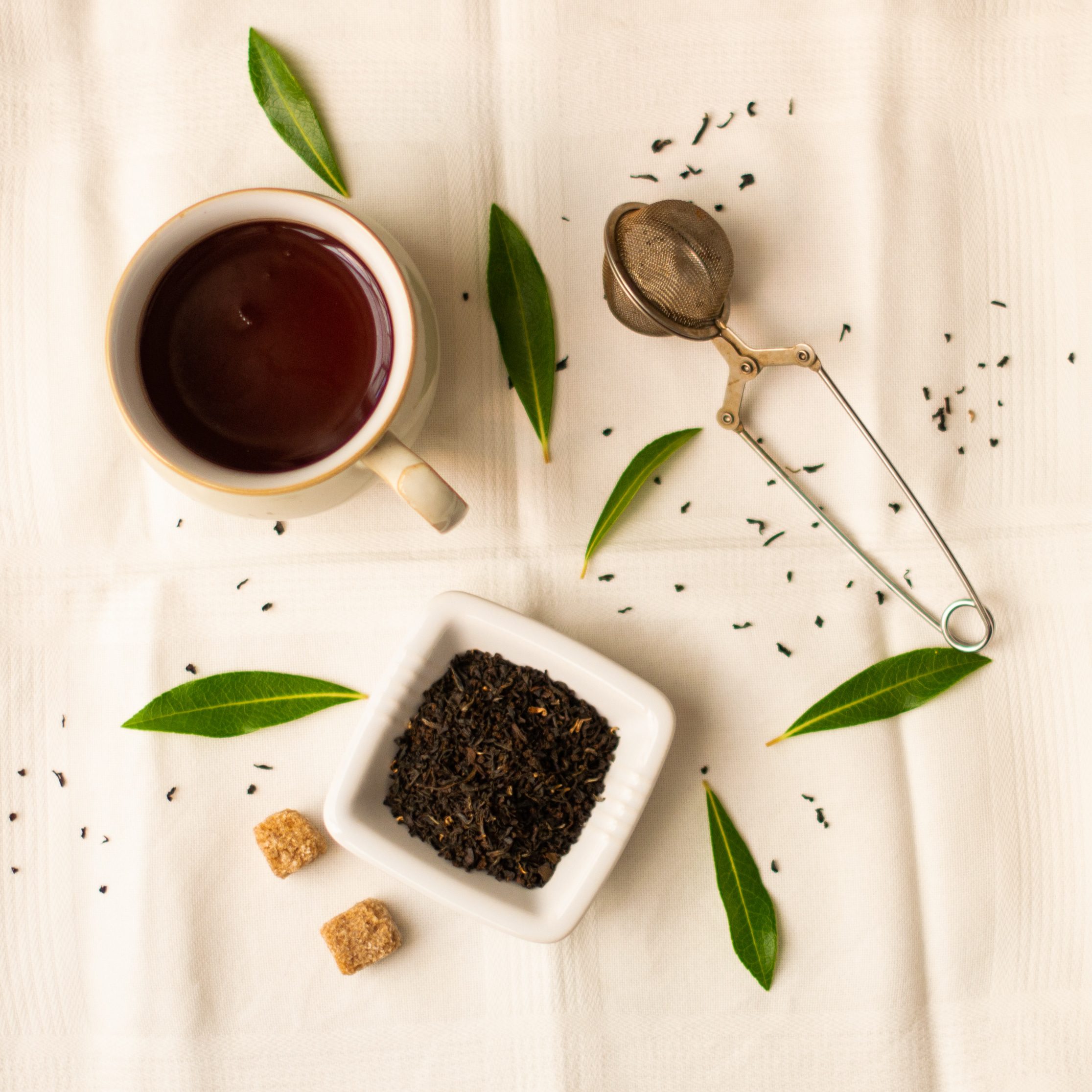 Cup of tea from above with green leaves, square dish of loose leaf tea and infuser 