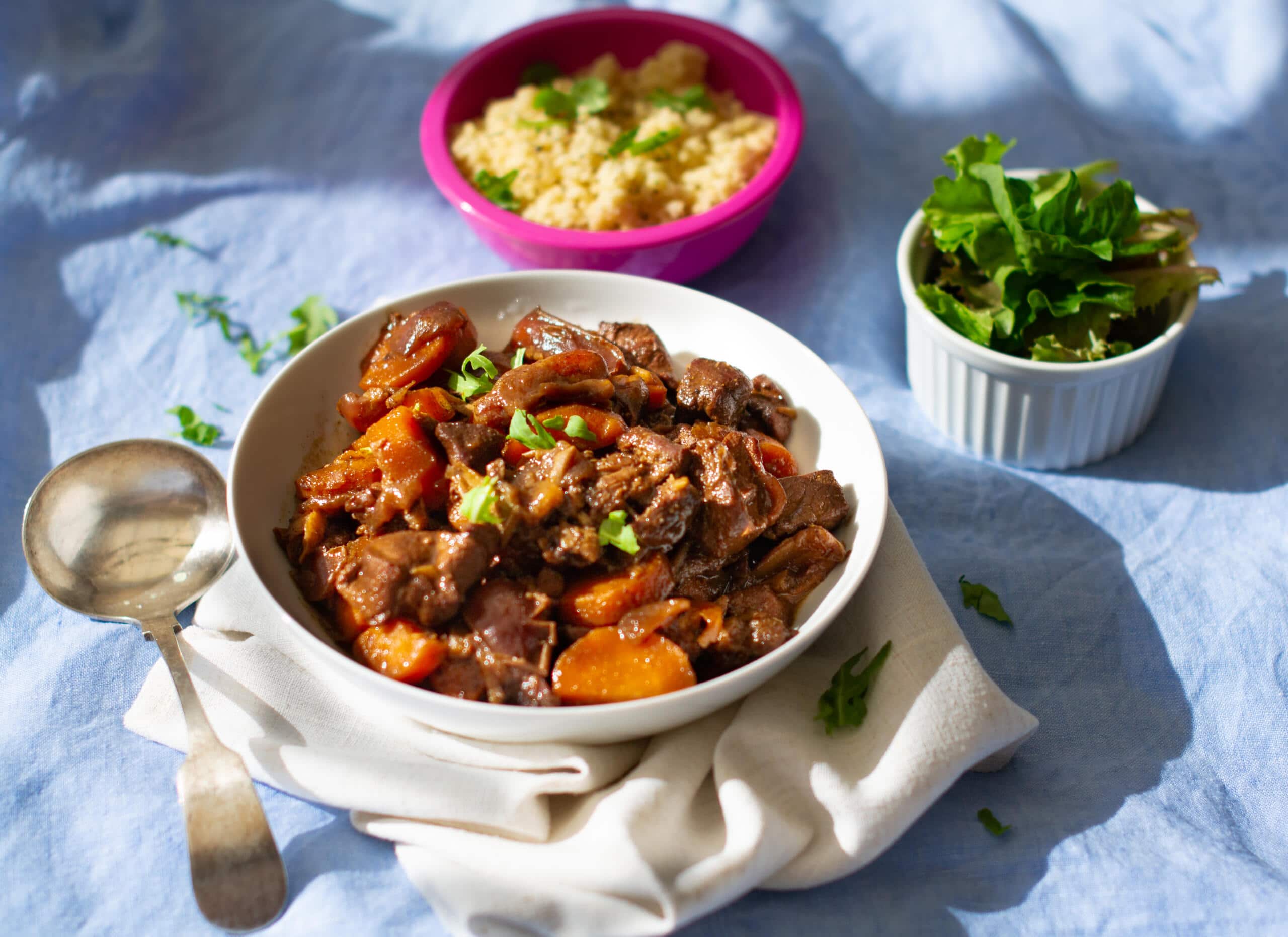 Bowl of lamb and coffee tagine on blue cloth served besides a pink bowl of cous cous and a white dish with salad
