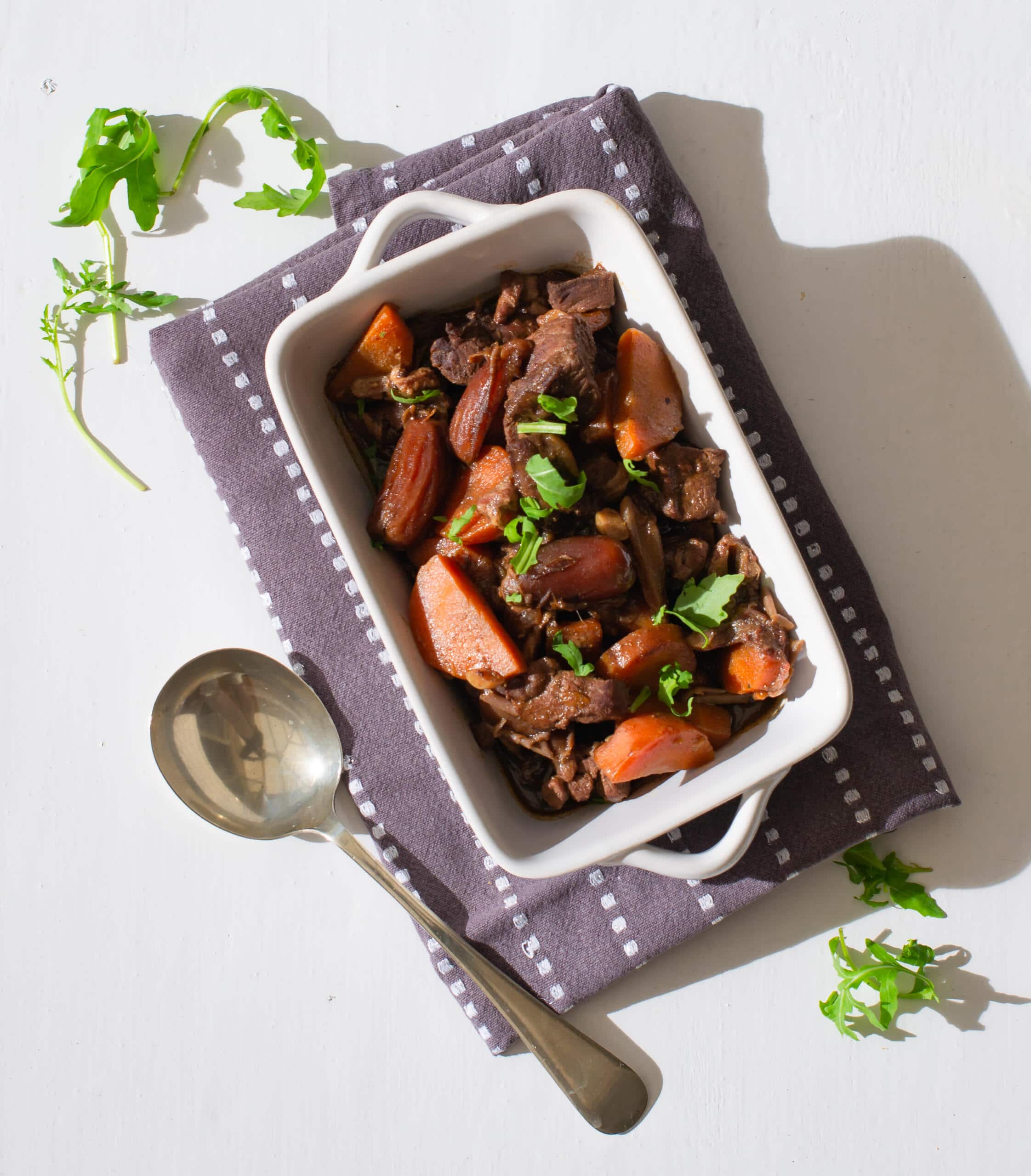 Oblong dish with lamb tagine placed on a dark grey tea towel with spoon and green leaves