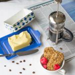 A cafetiere next to a a bowl of cereal, butter and a newspaper