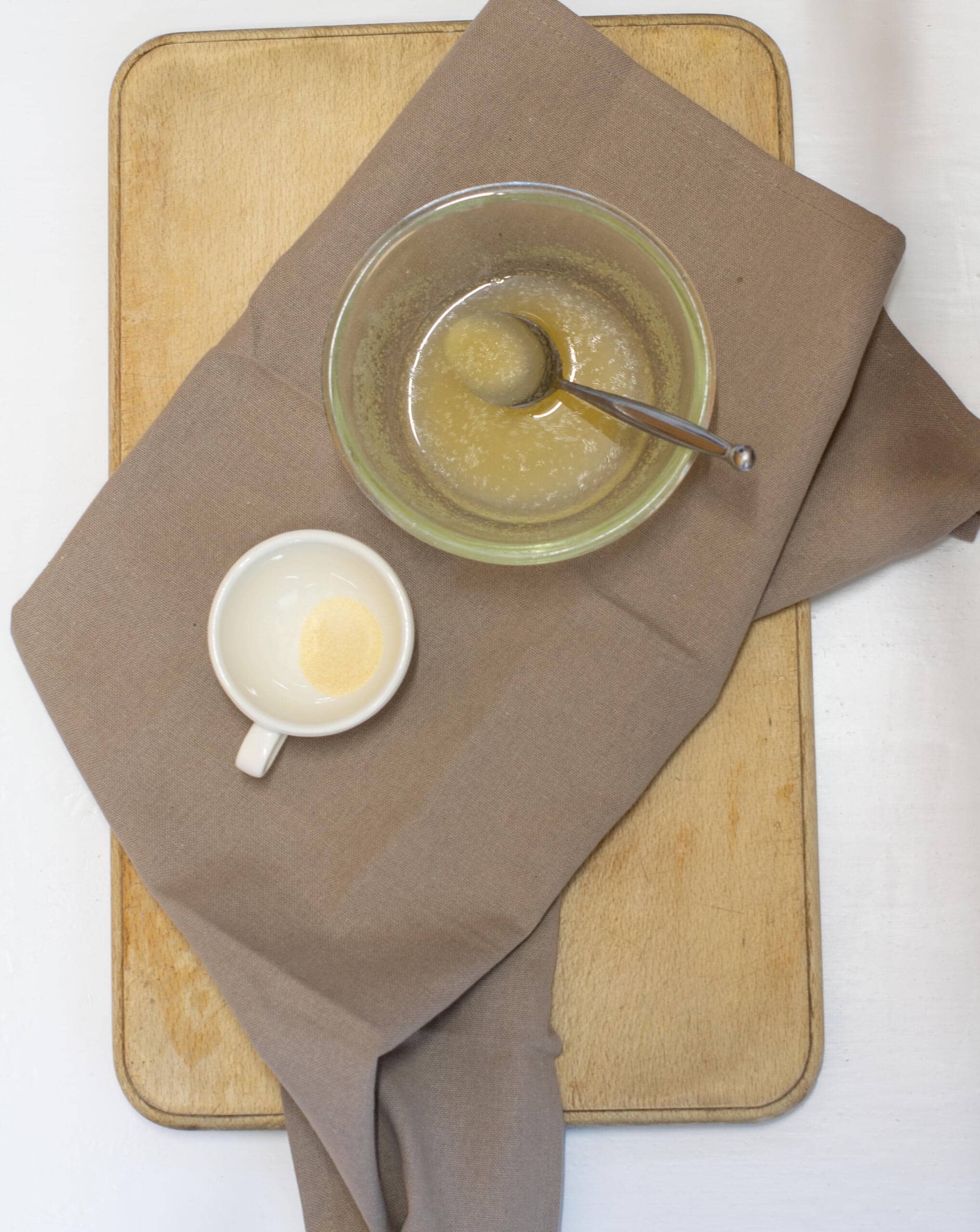 Glass bowl of powdered gelatine and water sitting on brown tea towel besides small cup