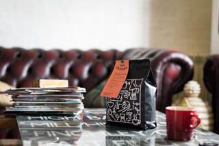 Dark roast Two Chimps coffee sitting on black table besides stack of magazines and red coffee mug