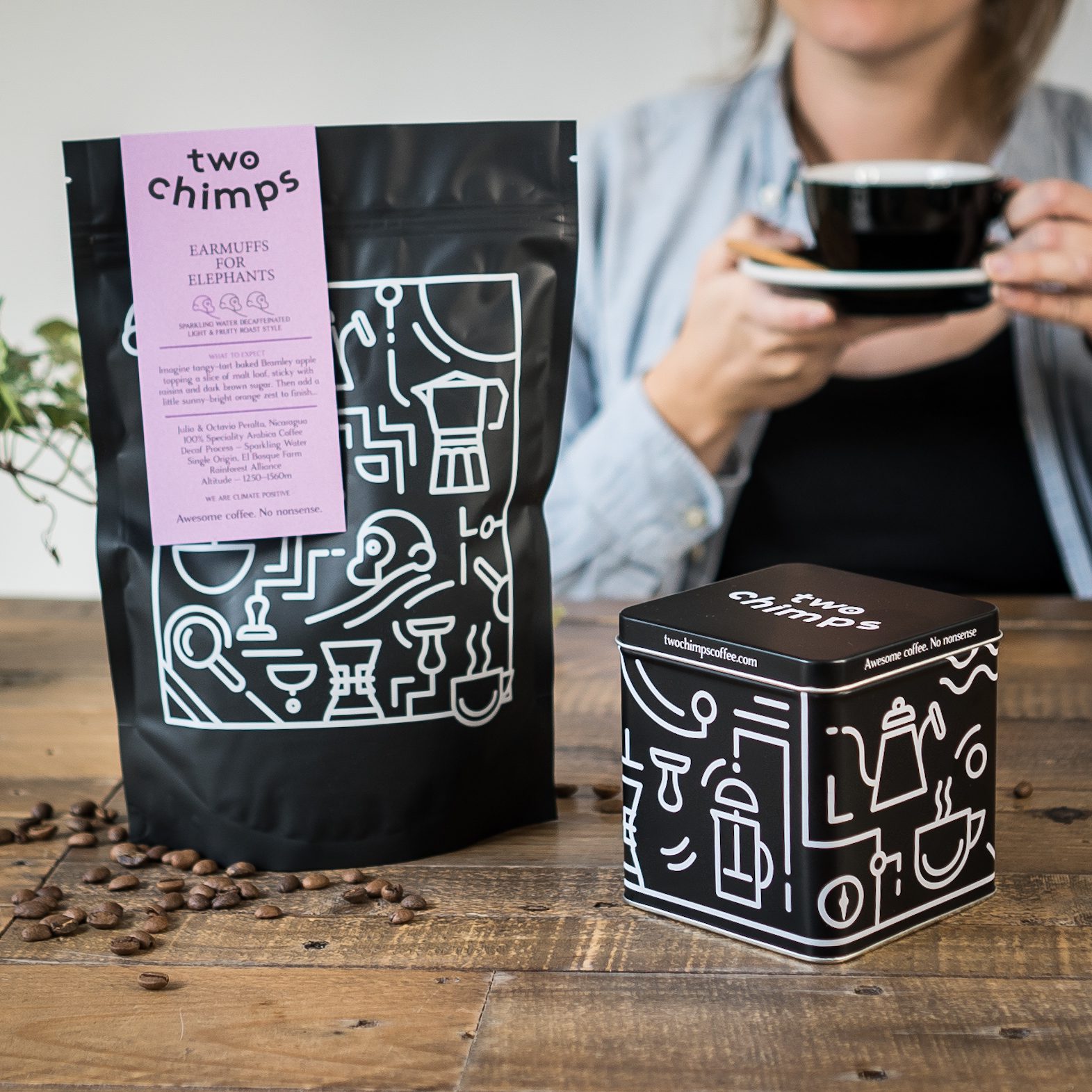 Bag of Two Chimps decaf coffee on a table with coffee tin and woman holding black cup and saucer