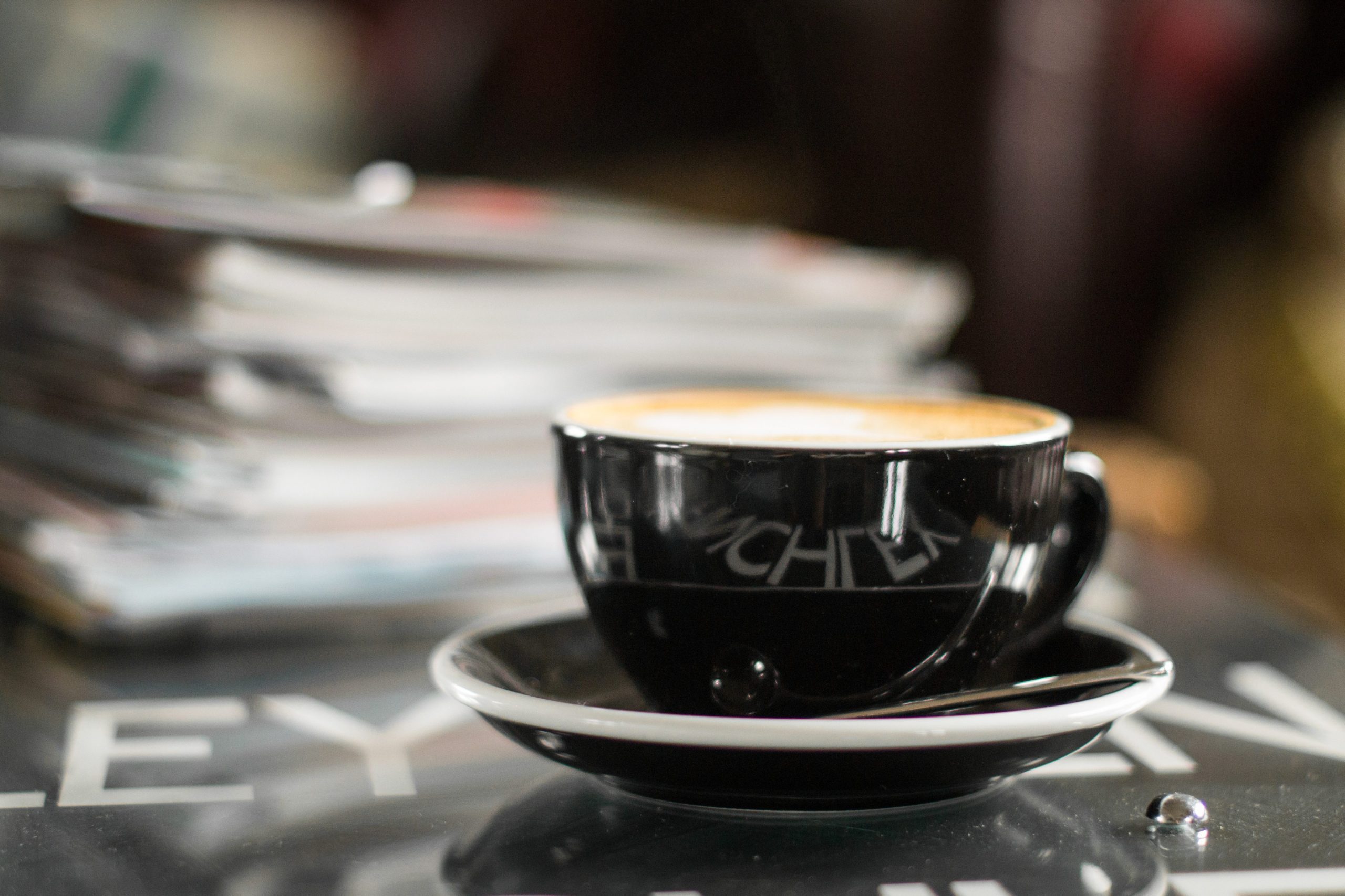Latte in a black cup and saucer beside stack of magazines